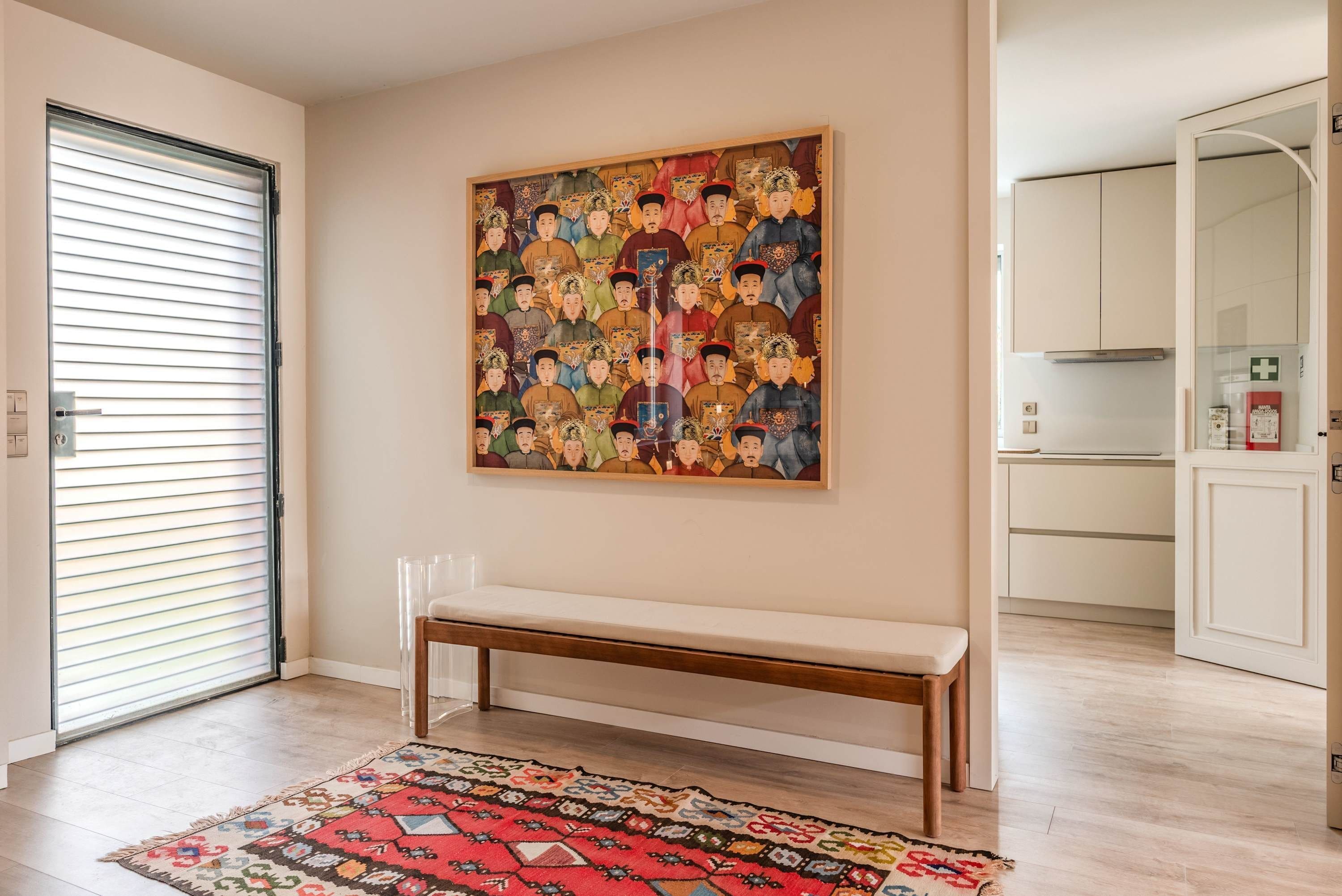 A room with a bench, a patterned rug, a large framed artwork, and a kitchen area.