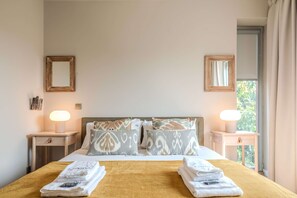 A hotel room with a bed, two bedside tables, two wall-mounted lamps, a window with curtains, and a view of greenery outside. - Liiiving in Porto - Foz Garden House (Porto)