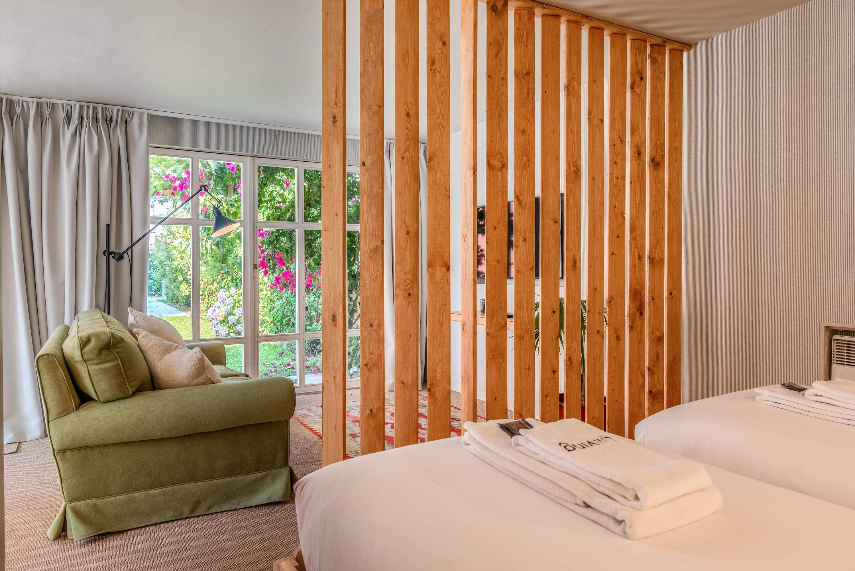 A hotel room with a green sofa, a bed with white linens, a wooden partition, and a window with a view of greenery and flowers.