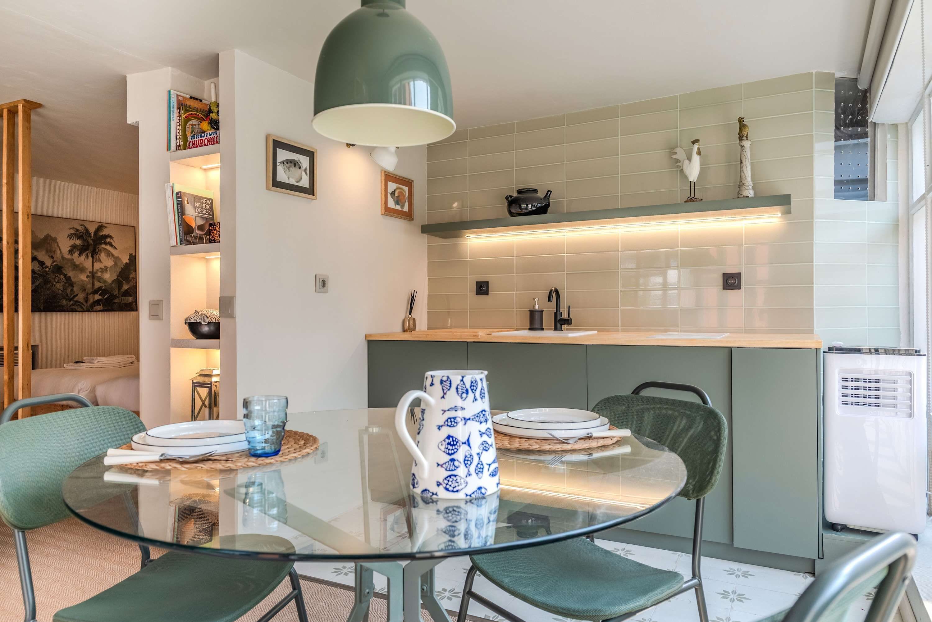 A modern kitchen with a glass dining table, green chairs, and a hanging light fixture.