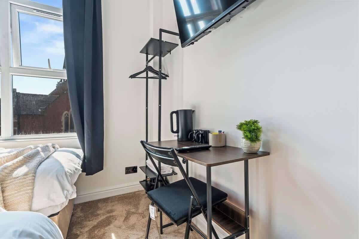 A small room with a bed, a desk with a chair, a television mounted on the wall, a window with curtains, and a potted plant.