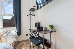 Ett litet rum med en säng, ett skrivbord med en stol, en tv som är monterad på väggen, ett fönster med gardiner och en krukväxt.