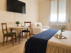 Ein Hotelzimmer mit Bett, Wandfernseher, Esstisch mit Stühlen, Heizkörper und Fenster mit Jalousien.