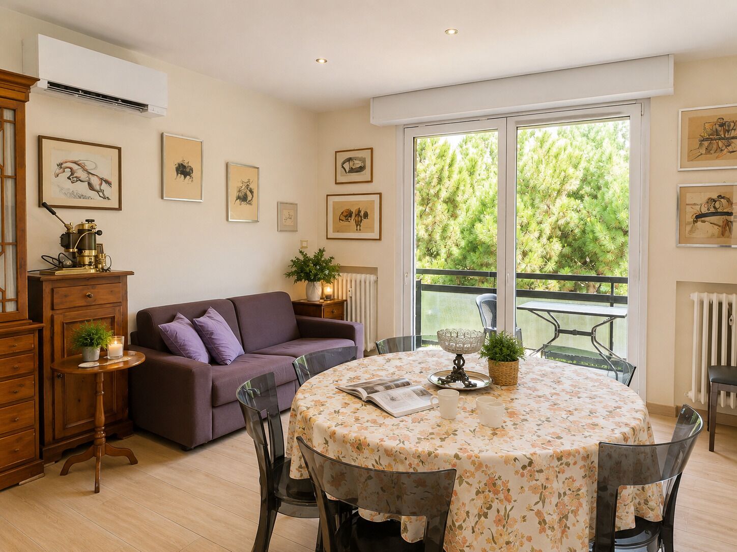 A dining area with a round table, chairs, a purple sofa, and a view of greenery through large windows.