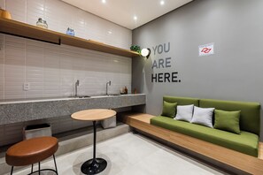 A modern kitchen with a built-in oven, a sink, and a countertop. There is a green sofa with cushions and a small round table with a stool. - StayHome Moema NexOne Pool & Gym (São Paulo)
