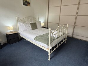 Un lit en métal blanc, doté d’une tête de lit en bois, se trouve dans une pièce où le sol est recouvert d’une moquette bleue. On y voit deux tables de chevet avec des lampes, ainsi qu’une étagère fixée au mur.