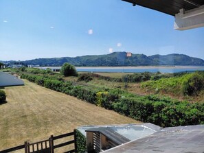 A view from a covered outdoor area with a lawn, a fence, and a body of water in the background. - Lovely Beach House In Deganwy | Conwy | Sleeps 6 (Deganwy)