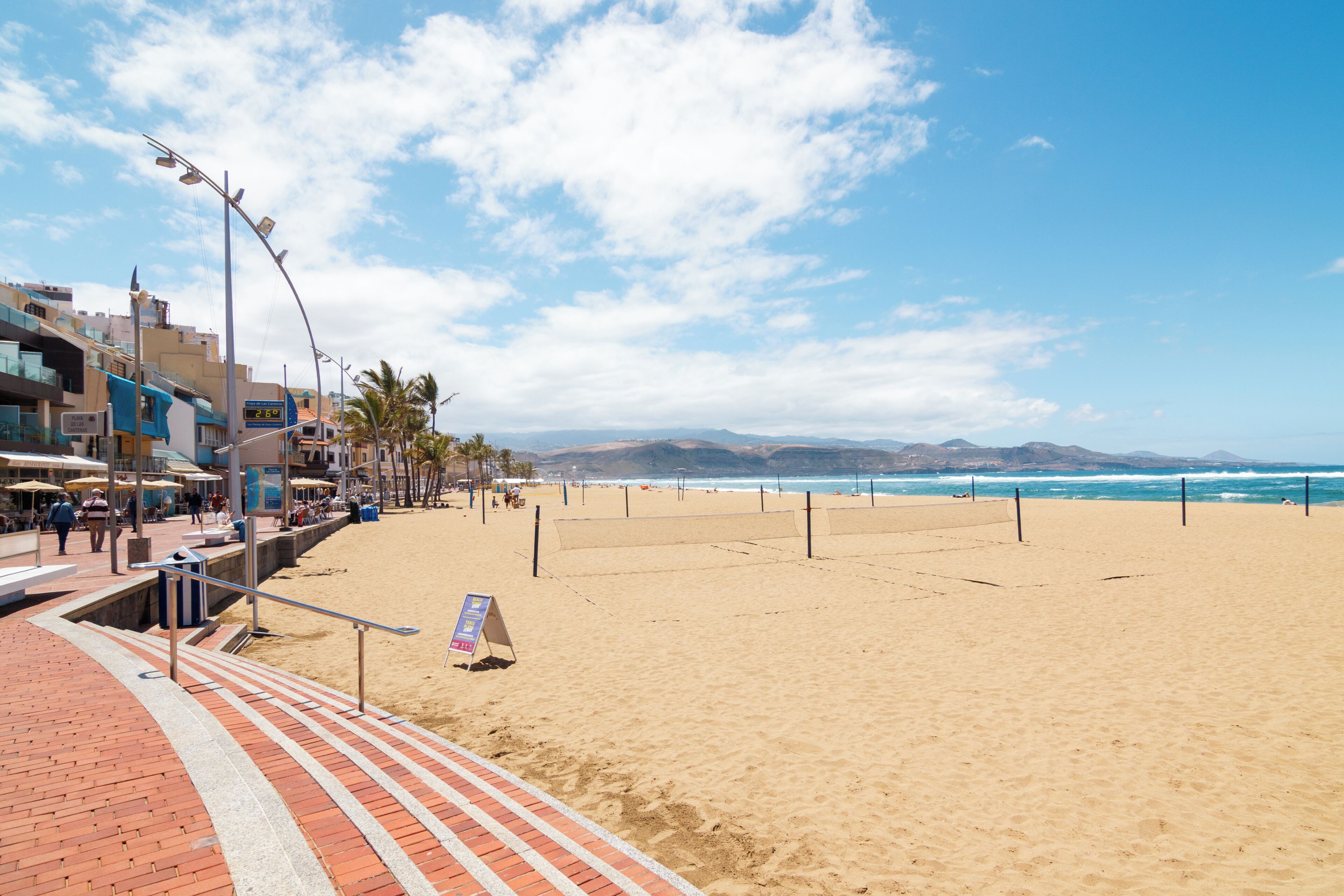 Una playa con un paseo marítimo, un cartel y edificios al fondo.
