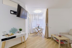 En moderne stue med et fladskærms-tv på væggen, et spiseområde med et glasbord og stole, samt en beige sofa.