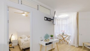 Una stanza moderna e luminosa con un letto, un comodino e una scrivania con una sedia. Si nota una ventola a soffitto, una televisione a parete e una finestra con tende leggere.