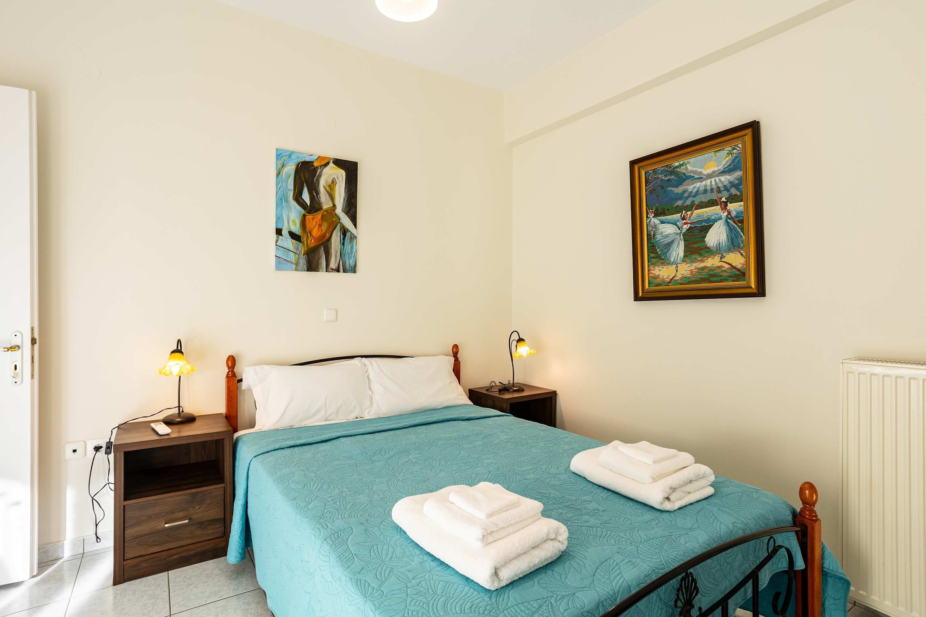 A bedroom with a bed, two bedside tables, a nightstand, a lamp, a painting, and a radiator.