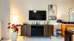 A modern living room with a flat-screen TV, a wooden cabinet, a white vase with flowers, and a small table with a lamp. - Kyparissia Modern Home Serene Views Calm Vibes (Trifylia)