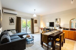 Ein stilvolles Wohnzimmer mit Sofa, Couchtisch, Esstisch und Stühlen. Es gibt einen Fernseher, einen Kamin und eine Küchenzeile mit Schränken und einem Fenster.