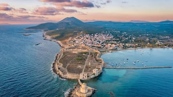 Vista aérea de uma vila costeira com uma estrutura fortificada, um farol e montanhas ao fundo.