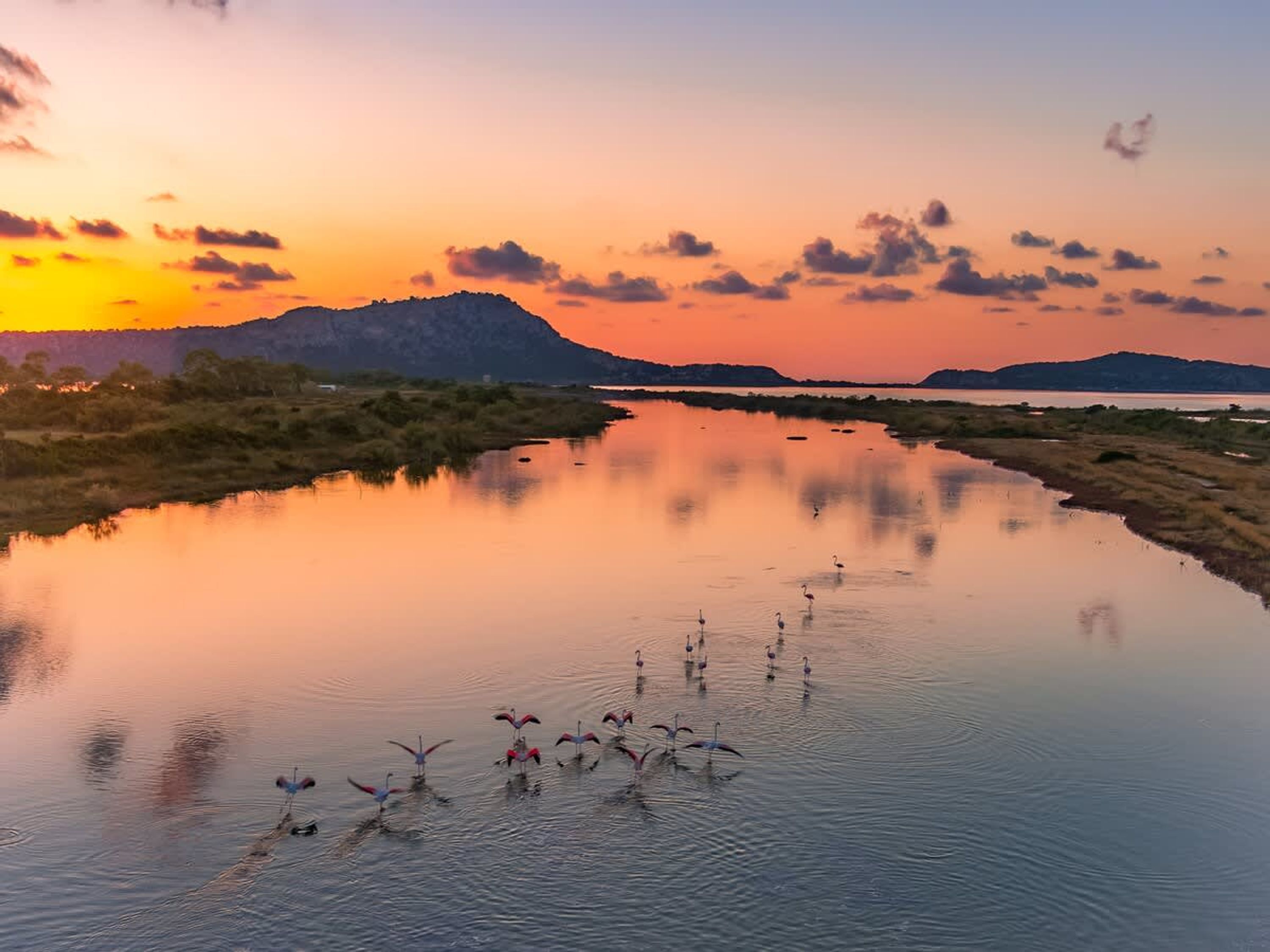 Água calma refletindo um pôr do sol, com uma montanha distante e vegetação escassa.