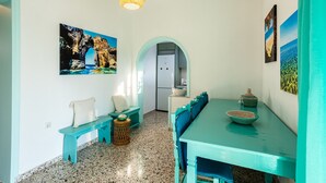 Un'area pranzo con un tavolo color petrolio, sedie e una panca. Sono presenti due quadri appesi al muro, una lampada in fibra naturale e una porta che dà su un'altra stanza.