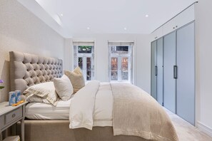 A bedroom with a tufted headboard, a bed with white bedding, a nightstand with a lamp, and a wardrobe. - Garden Apartment Escape in Hampstead (London)
