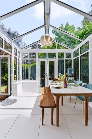 A conservatory with a dining table, benches, and a hanging light fixture. - Garden Apartment Escape in Hampstead (London)