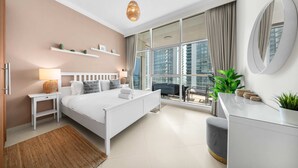 Un dormitorio moderno con cama, mesitas de noche, cómoda y un amplio ventanal con cortinas, que ofrece vistas de edificios altos.