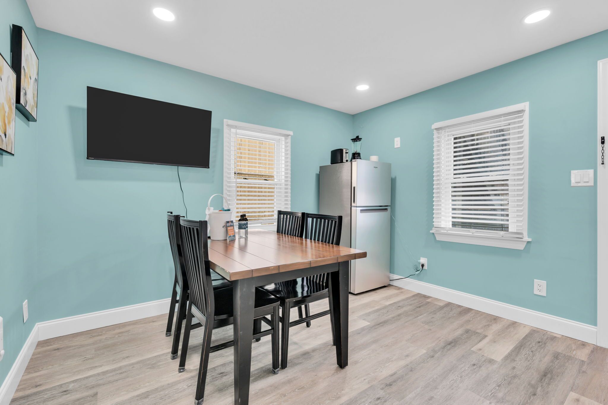 A dining area with a table and chairs, a flat-screen TV mounted on the wall, a refrigerator, and a window with blinds.