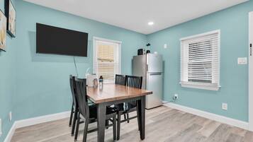 A dining area with a table and chairs, a flat-screen TV mounted on the wall, a refrigerator, and a window with blinds.