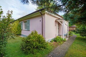 Ein eingeschossiges Haus mit einer kleinen Veranda, umgeben von Grünpflanzen und einem gepflasterten Weg.