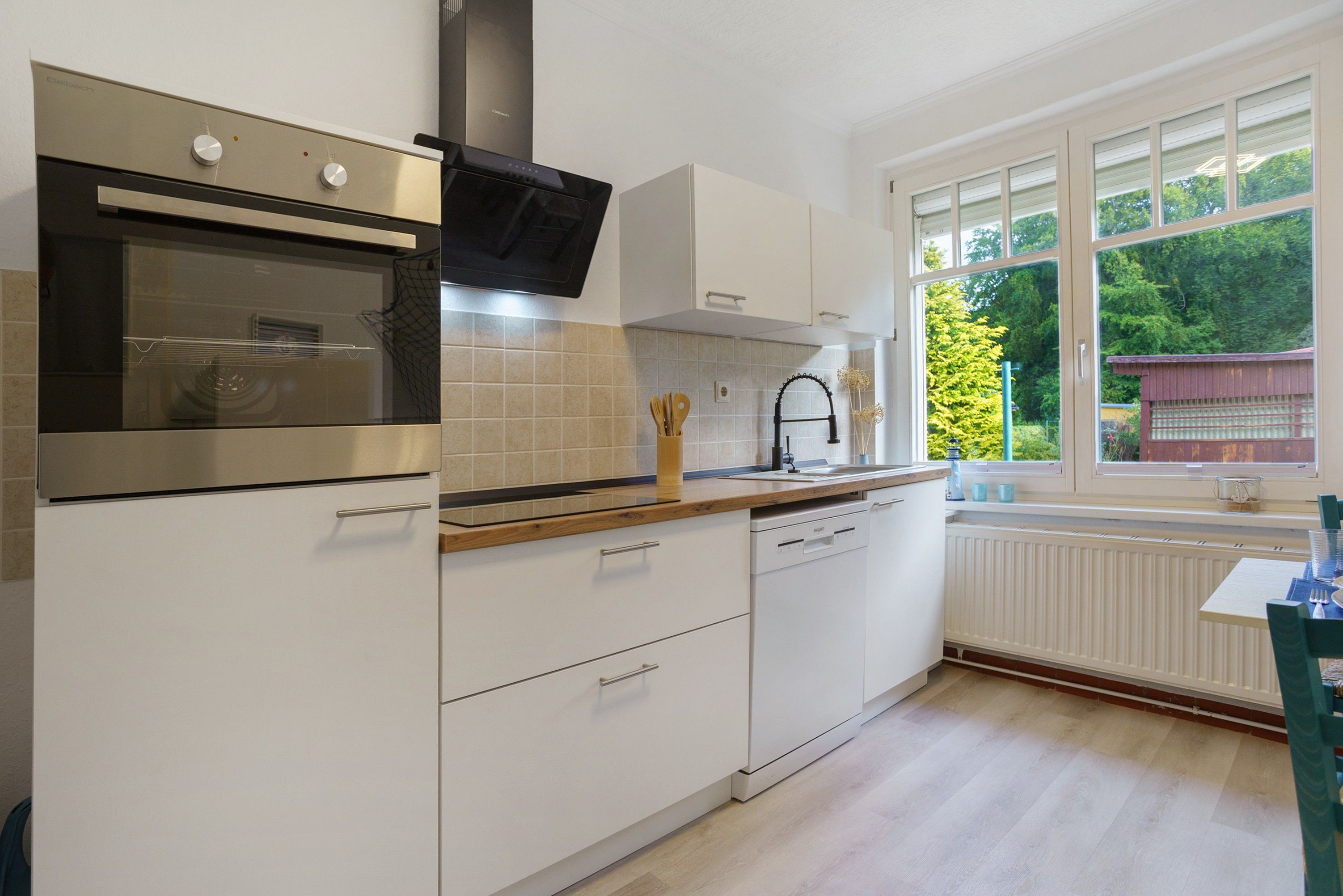 A modern kitchen with a built-in oven, a dishwasher, and a window with a view of greenery.