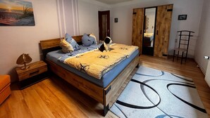 A bedroom with a wooden bed, a nightstand, a lamp, a chair, and a view of a hallway. - Apartment 'Idylle Ostsee' – Buddenhagen, Gateway to Usedom Island (Wolgast)