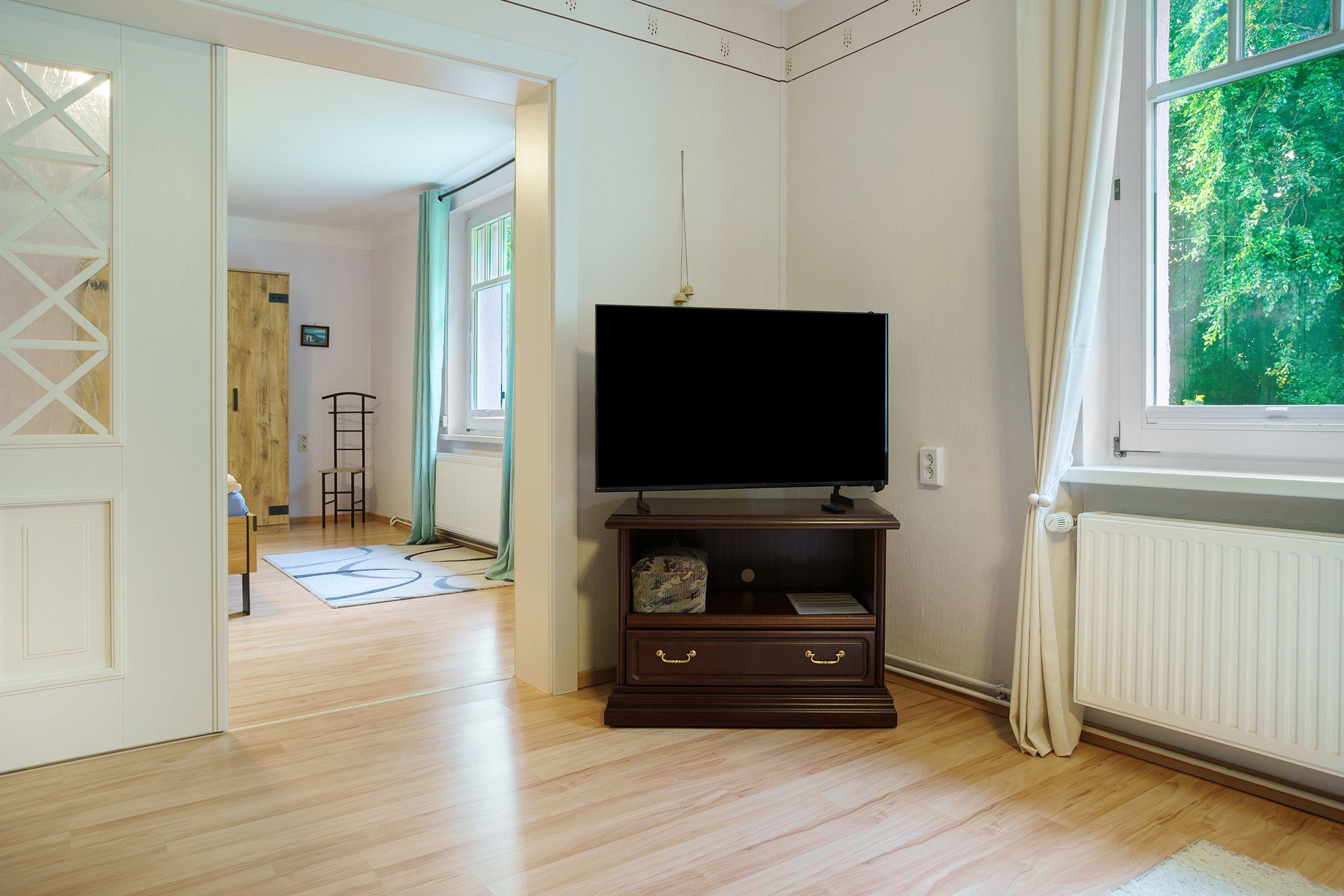 A room with a television on a wooden stand, a radiator, a window with curtains, and a wooden floor.