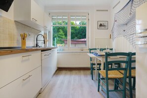 A kitchen with white cabinets, a window with a view of greenery, and a dining area with a table and chairs. - Apartment 'Idylle Ostsee' – Buddenhagen, Gateway to Usedom Island (Wolgast)