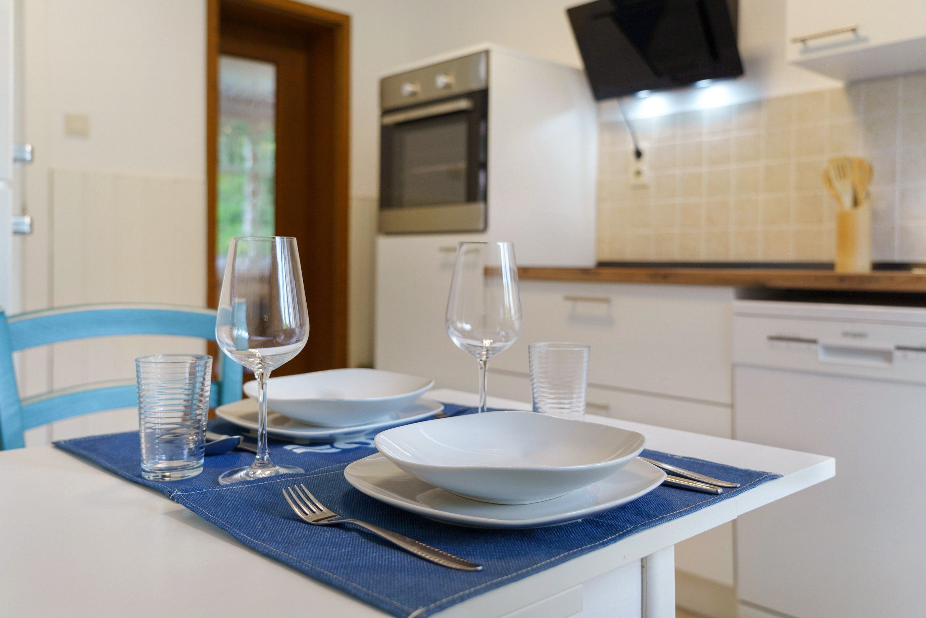 A dining table set with plates, glasses, and cutlery, with a kitchen in the background.