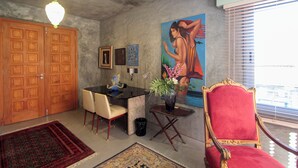 Een kamer met een eettafel, stoelen, een rode fauteuil, een schilderij en een groot raam met jaloezieën.