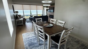 Un coin repas avec une table en bois et des chaises blanches, une vue sur la plage et une grande fenêtre.