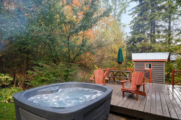 A hot tub on a wooden deck with outdoor seating and a small cabin in the background.