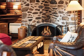 A stone fireplace with a fire, a wooden table with books, a leather chair, and a lamp.