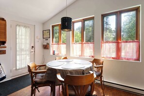 A dining area with a round table, chairs, a hanging light fixture, and large windows with curtains.