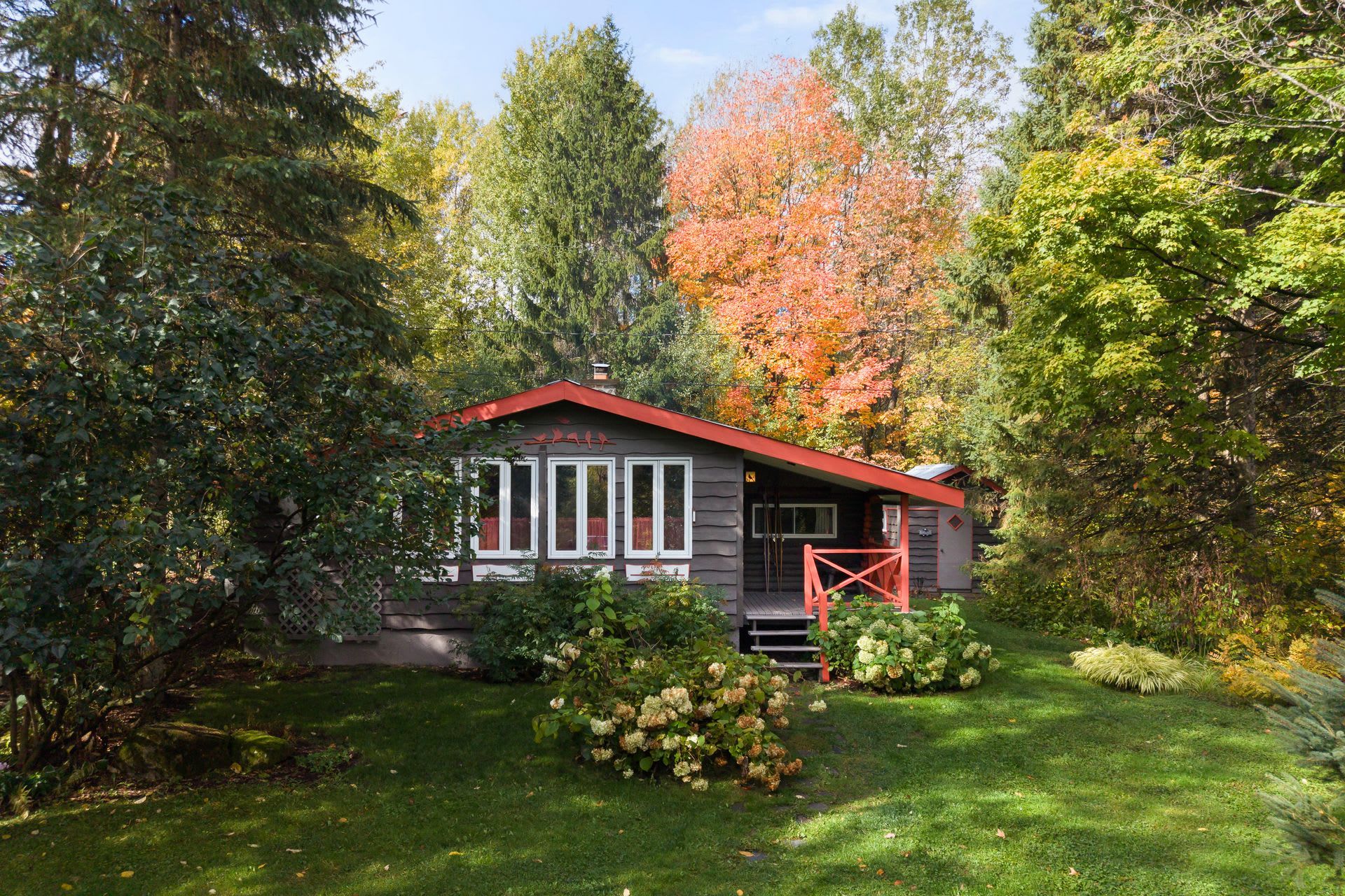 Une petite cabane avec un toit rouge et des boiseries blanches, entourée d’une végétation luxuriante et d’arbres.