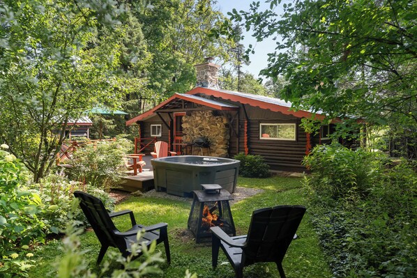 A rustic cabin with a stone exterior, a hot tub, and a fire pit in a lush garden setting.