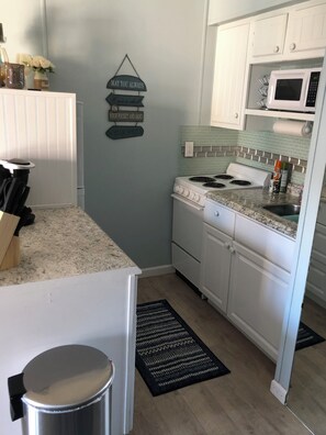 A compact kitchen with white cabinets, a stove, a microwave, and a trash can. - Cozy 1-bedroom condo in serene Corpus Christi. (Corpus Christi)