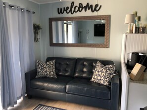A living room with a black leather sofa, patterned cushions, a 'welcome' sign, a mirror, and a coffee machine. - Cozy 1-bedroom condo in serene Corpus Christi. (Corpus Christi)