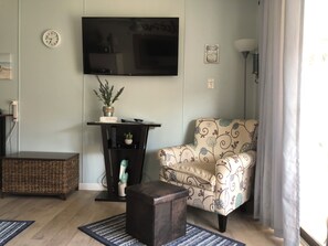 A living room with a TV mounted on the wall, a decorative plant, a patterned armchair, a small wooden table, and a wicker side table. - Cozy 1-bedroom condo in serene Corpus Christi. (Corpus Christi)