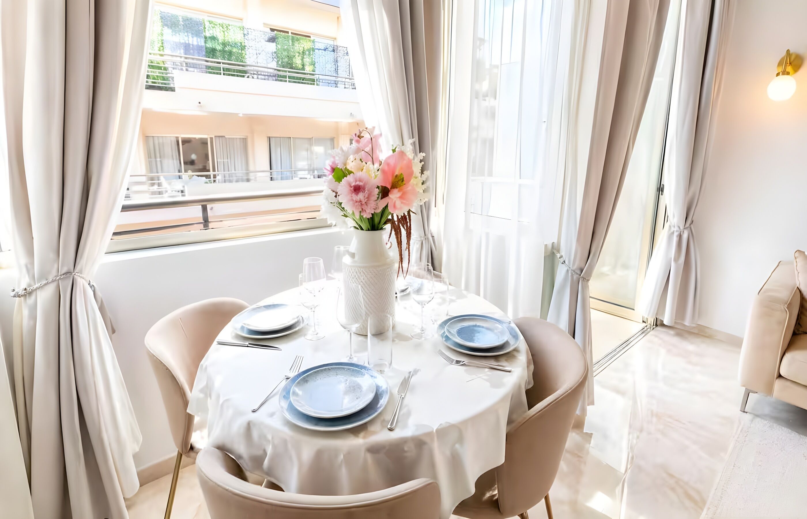 A dining table set with plates, glasses, and a vase of flowers, with a view of a balcony and building outside.