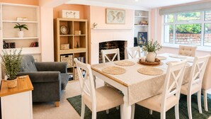 A dining area with a table set for four, surrounded by white chairs. There is a bookshelf with various items and a potted plant on a side table. - The Willow Chester | Parking | Canal Walks | Large Garden | Sleeps 8 (Chester)