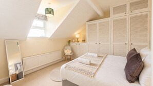A bedroom with a sloped ceiling, a bed with pillows, a chair, a mirror, and a wardrobe. - The Willow Chester | Parking | Canal Walks | Large Garden | Sleeps 8 (Chester)