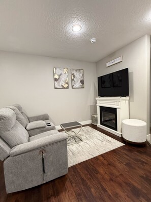 Ein Wohnzimmer mit einem grauen Ecksofa, einem Kamin, einem Flachbildfernseher und zwei abstrakten Wandbildern.