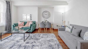 Ein modernes Wohnzimmer mit einem Glastisch, einem grauen Sofa, einem grünen Sofa, einem Essbereich mit Tisch und Stühlen sowie einer Wanduhr.