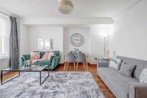 A modern living room with a glass coffee table, a grey sofa, a green sofa, a dining area with a table and chairs, and a wall clock. - Fantastic 1 Bedroom Apartment Just Off Marble Arch (London)