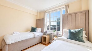 A hotel room with two beds, a window with a city view, and a small wooden side table. - Beautiful 2 Bedroom off Marble Arch (London)