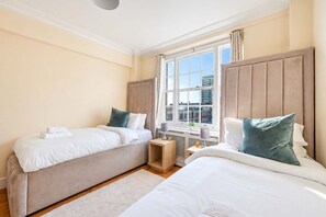 A hotel room with two beds, a window with a city view, and a small wooden side table. - Beautiful 2 Bedroom off Marble Arch (London)