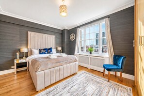 A bedroom with a large bed, a bedside table, a chair, a window with curtains, and a clock on the wall. - Beautiful 2 Bedroom off Marble Arch (London)